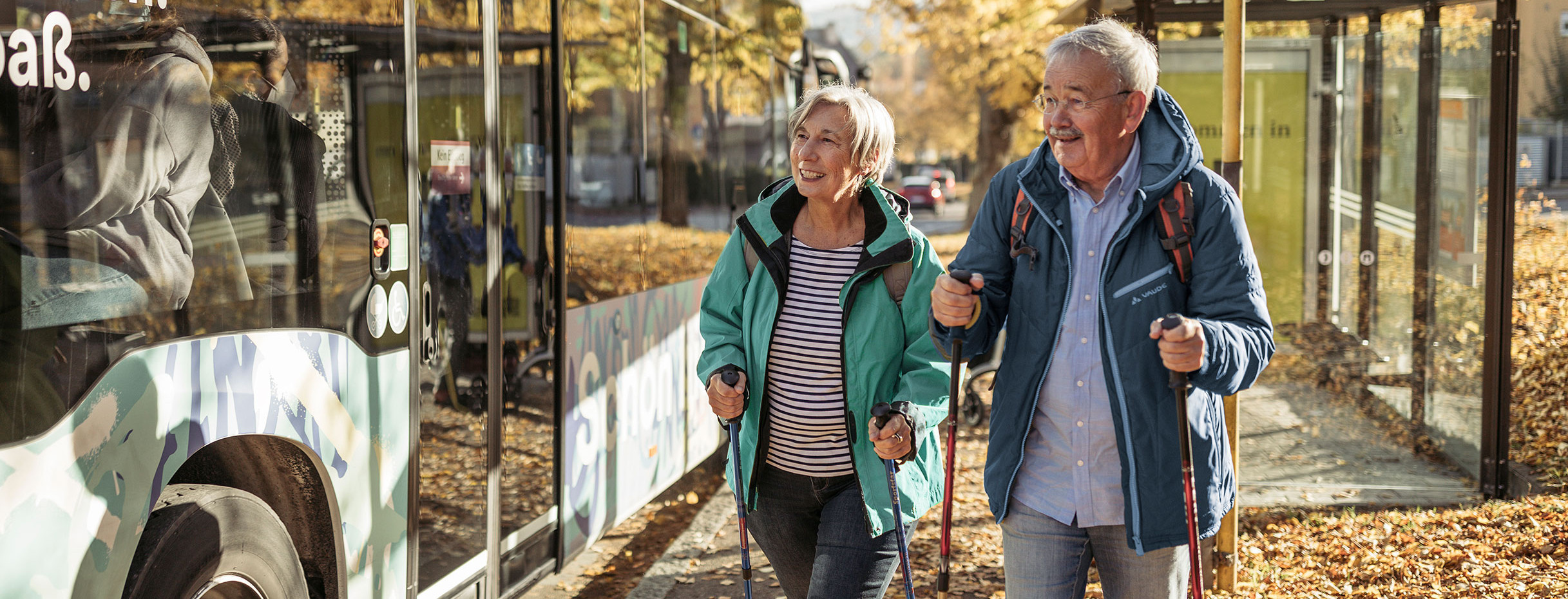 Zwei ältere Menschen in Wanderkleidung laufen an einer Bushaltestelle vorbei.